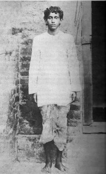 Full-length photograph of Khudiram Bose as a young man, standing barefoot in a long kurta and dhoti against a brick wall.